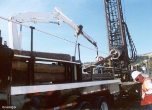 The crane is supporting the sampler barrel while it is being connected to the vibratory drill head. Notice how the drill head is rotated on the mast to facilitate the quick connecting and disconnecting of sampler barrels and drill stem.