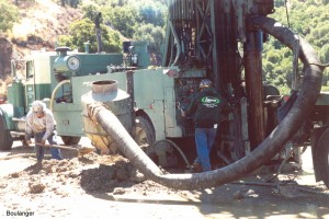  During open-bit drilling, the return air carries the soil cuttings to a "cyclone" where the soil loses velocity and drops out the bottom onto a cuttings pile. A large flexible tube carries the air return from the top of the casing (upper-right of photo) to the cyclone (mid-left of photo). The field engineer must carefully record the nature of the cuttings, the diesel hammer's bounce chamber pressure, and the hammer-blow rate throughout drilling.
