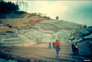 This is view of the exposed bedrock on the right abutment of the dam. The people in the foreground are standing on compacted shell material. The rock of the foundation and abutments must be prepared before compacted soil can be placed against it, and the dam raised to its final elevation. The granitic gneiss rock has a very uneven surface due to weathering along joints and layering.