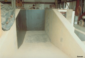 The instrumented wall, on the right, consists of four concrete panels, each 2.5 feet wide by 7 feet high. The panels are supported on load cells which measure the horizontal and vertical load on each panel. Earth pressure cells are embedded flush with the faces of the middle two panels. The black walls at the left and on the far end are designed to reduce wall friction.