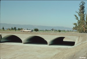 Here three culverts, side-by-side, provide a bridge across a river. These are “arch culverts,” without metal inverts. The haunches of the metal arches are supported on concrete footings, and a concrete invert prevents erosion of the stream bed in periods of high flow in the river.