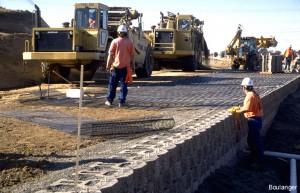 Geogrids are being laid out over a completed row of blocks. The two scrapers (earth-moving equipment in the upper left) are placing fill soils behind the geogrids.