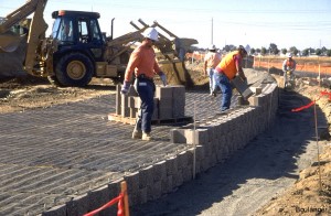 Another row of blocks is being placed, with the geogrid sandwiched between the overlapping blocks. The front-end loader is placing the drainage layer in the background.