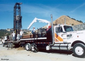 The sonic drilling rig is in the background. The truck in the foreground is equipped with a small crane for handling the large sampler barrels and drill stems.